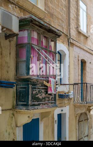 A traditional Maltese balcony, adorned with pink curtains and laundry drying on a line, adds character to the aged facade of a building in Malta. Stock Photo