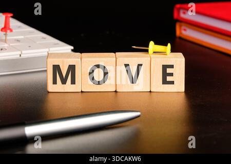 Wooden blocks spell MOVE on a dark desk next to a pen. Push pins, a white keyboard, and notebooks are nearby, hinting at action, change, or relocation Stock Photo