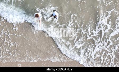 Happy couple enjoying a playful walk on the beach, laughing and holding hands as they splash through the waves together || Model released Stock Photo