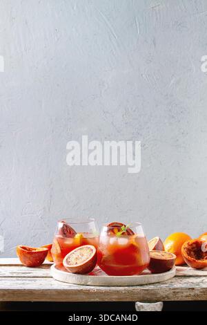 Blood orange iced cocktails in glasses, decorated by slice of oranges and fresh mint, served in ceramic tray on white wooden table with grey wall at background. Stock Photo