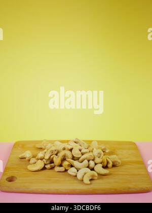 Small pile of raw cashew nuts placed on a wooden board against a bright yellow background, clean minimal ingredient presentation for healthy snack Stock Photo