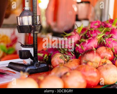 A juice press is used to extract juice from pomegranates while dragon fruits are arranged on an orange stand. Fresh fruit is the focus of this market Stock Photo