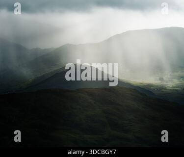 Sunlight shines through the clouds and rain, casting strong shafts of light and shadow on the mountains. Hills and valleys stretch into the distance. Stock Photo