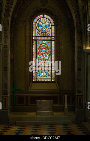 Rich stained glass fills a dim church interior, framed by decorative arches and tiled surfaces. A stone altar sits in the foreground, inviting reflect Stock Photo