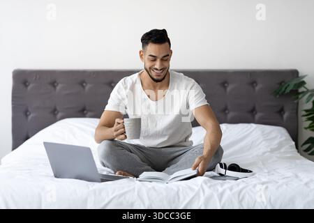 Young Arab Man Enjoying Freelance Work in Bed With Coffee, Laptop, and Notepad Stock Photo