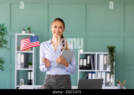 Happy Foreign Language Teacher Holding a Tablet Next to a Desk With a USA Flag Stock Photo