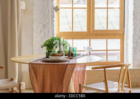 Minimalist Christmas dining table styled in Scandinavian interior on wide window background with natural textiles, greenery, candles. Festive home atm Stock Photo
