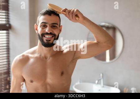Young Man Brushes Hair While Smiling in Modern Bathroom at Home Stock Photo