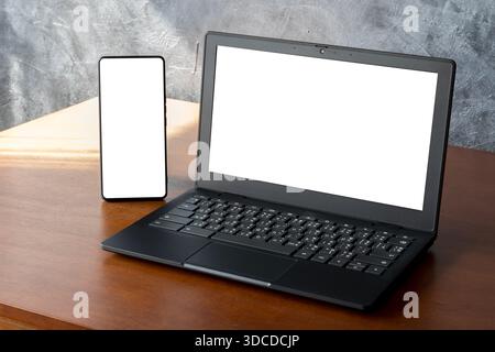 Close-up of black laptop and smartphone with blank white screens on wooden desk, concrete wall background and sunlight. Ideal for UI mockups, ads, rem Stock Photo
