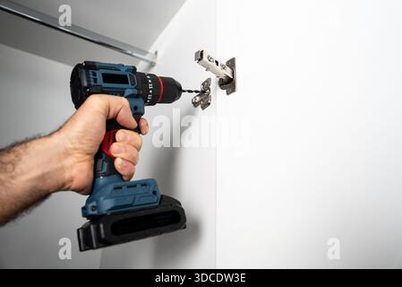 Close-up of a handyman installing a metal hinge on a wall using a cordless drill. Home renovation and furniture mounting concept. Stock Photo