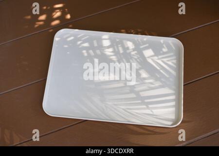High angle view of white glossy rectangular tray empty with shadow pattern light texture on brown wooden floor background creating calm peaceful atmos Stock Photo