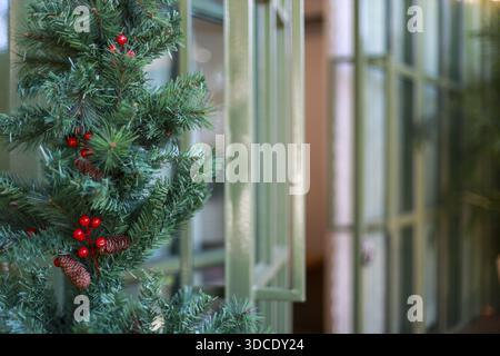 Christmas decoration green pine tree branch with red berry on blurred window background creates festive holiday mood and winter season atmosphere for Stock Photo