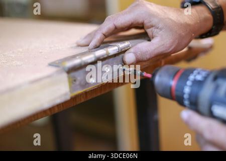 Concentrated carpenter installing door hinge carefully while using cordless electric screwdriver drill on plywood board with professional attention to Stock Photo