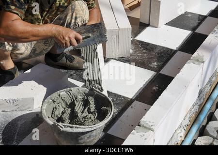 Skilled male worker hands holding trowel carefully applying grey cement mortar from bucket installing durable black white granite tile on outdoor floo Stock Photo