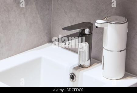 Close-up of a chrome faucet and white touchless liquid soap dispenser in a minimalist bathroom interior. The concept of hygiene, cleanliness. Stock Photo
