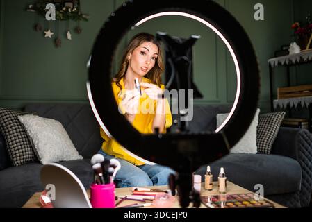 Beauty vlogger filming a makeup tutorial at home. She is sitting on sofa and talking about lipstick. Stock Photo