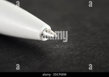 Detailed view of the metallic ballpoint tip of a white pen resting on a dark, slightly gritty background, highlighting fine textures and subtle ink re Stock Photo