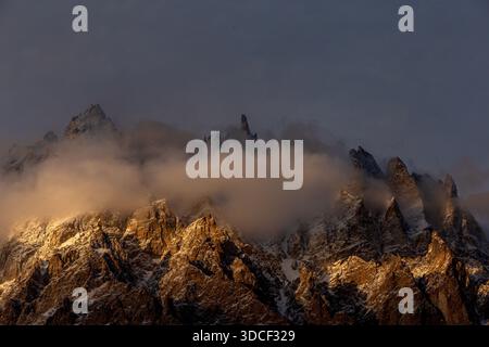 View of jagged, snow-dusted peaks pierce through swirling clouds, bathed in the warm glow of sunset along the Karakoram Highway, Passu, Gilgit Baltist Stock Photo