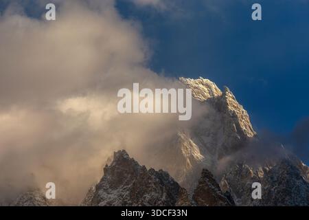 View of jagged, snow-dusted peaks piercing through a swirling veil of clouds under a crisp, azure sky, bathed in golden sunlight, Passu, Gilgit Baltis Stock Photo