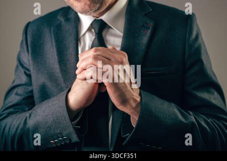 Businessman in dark suit with hands firmly clasped together in front of his chest, conveying intense worry, hope, prayer, or desperation. Selective fo Stock Photo