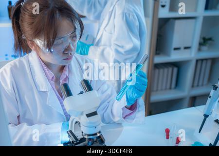 Two female scientists in white lab coats working together in a bright laboratory with microscopes, glassware, and blue test tubes, representing teamwo Stock Photo