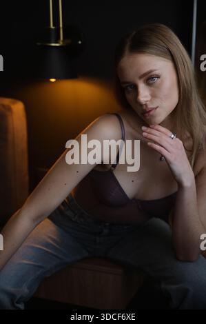 Slender blonde woman with long hair in a mauve bodysuit and jeans on a solid dark background Stock Photo