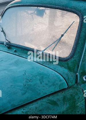 Old green car covered in frost. Close-up of frosted windshield and hood on a classic vehicle, warm morning light illuminates the cabin from behind. Stock Photo