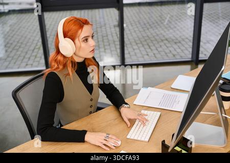 Young woman with red hair attentively works at her desk while wearing headphones in a bright office. Stock Photo