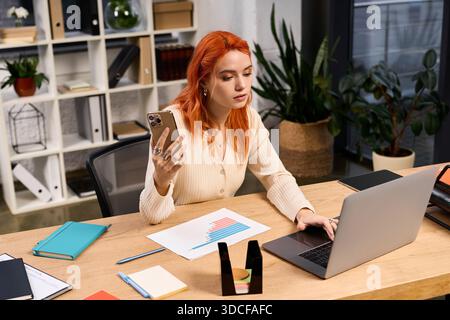 Young woman works attentively at her desk, using a laptop and smartphone in a sleek office. Stock Photo