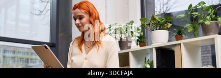 A young woman with red hair uses her tablet in a modern, green-filled office, banner Stock Photo