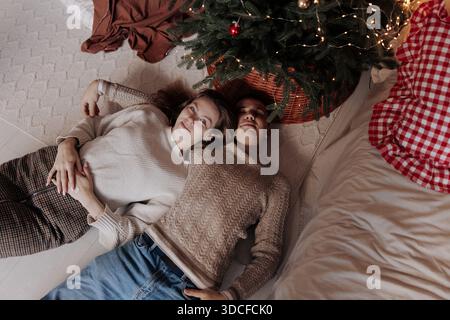 Top view of a man and woman relaxing together on a rug near a decorated Christmas tree with lights. They are looking up at the camera, enjoying a peac Stock Photo