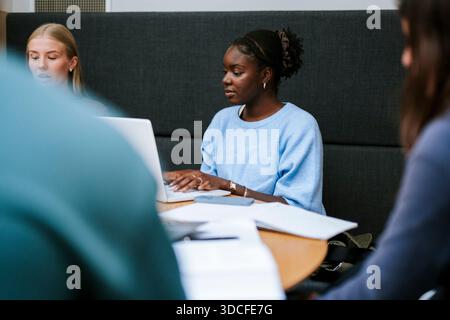 Concentrated young woman using laptop while sitting with friends in university campus Stock Photo