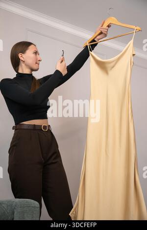 Fashion designer taking pictures of a beige silk dress hanging on a wooden hanger with her smartphone, creating visual content for online store or soc Stock Photo