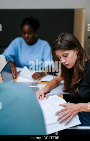 Young female student reading book while studying with friends at college campus Stock Photo