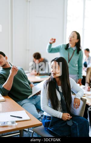 Long haired young female student listening to teacher while sitting at desk in classroom at college Stock Photo