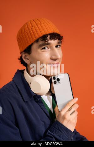 Stylish man in an orange hat holds a phone and wears headphones against a warm backdrop. Stock Photo