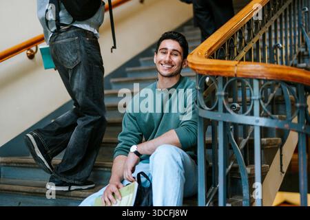 Smiling portrait of young male student sitting on stairs in college building Stock Photo