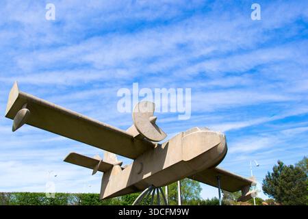 View of a stone-like airplane sculpture against a bright blue sky with wispy clouds, grounded on a metal stand in Biscarosse, France. Stock Photo