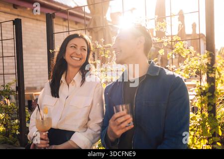 Smiling man and woman looking at each other while sitting outdoors with drinks at sunny day Stock Photo