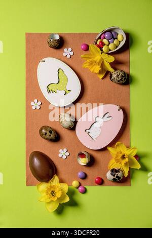 Easter greeting card with wooden decorations, chocolate sweets and eggs, daffodils on bright felt orange green background. Flat lay, copy space. Stock Photo