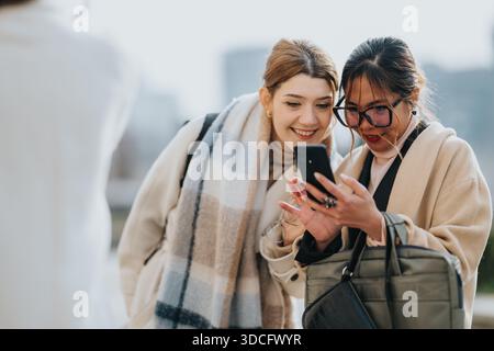 Two business women look at a smartphone together outdoors in the city, sharing ideas and smiles Stock Photo