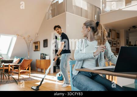 Young woman sitting with laptop while looking at boyfriend vacuuming floor in home Stock Photo
