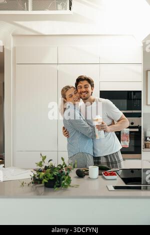 Smiling young man embracing girlfriend while standing in kitchen at home Stock Photo