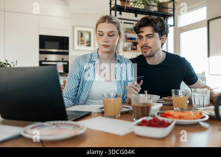 Young couple checking bank statement on laptop while having breakfast on dining table at home Stock Photo