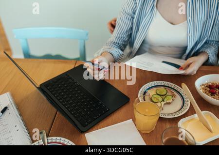 High angle view of young woman holding credit card while sitting with laptop on dining table in home Stock Photo