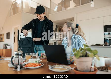 Smiling young man keeping laptop in backpack near girlfriend sitting at table in home Stock Photo