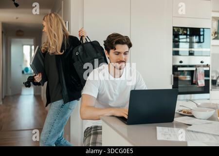 Young man using laptop at home while girlfriend leaving for work Stock Photo