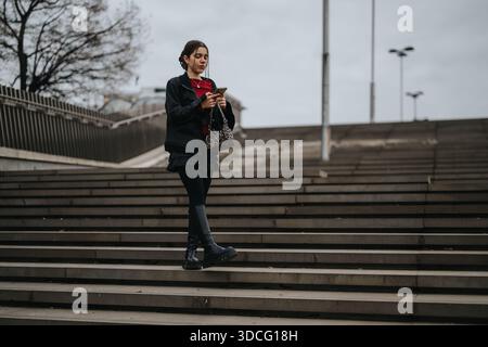 Young woman on urban stairs using smartphone, stylish jacket and boots, overcast day, alone outside Stock Photo