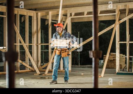 A worker holds wooden planks at a construction site with wooden frames in place. Tools are visible on his belt. Stock Photo