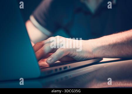 A person types on a laptop with one hand while sitting at a table. The room is dimly lit. Stock Photo